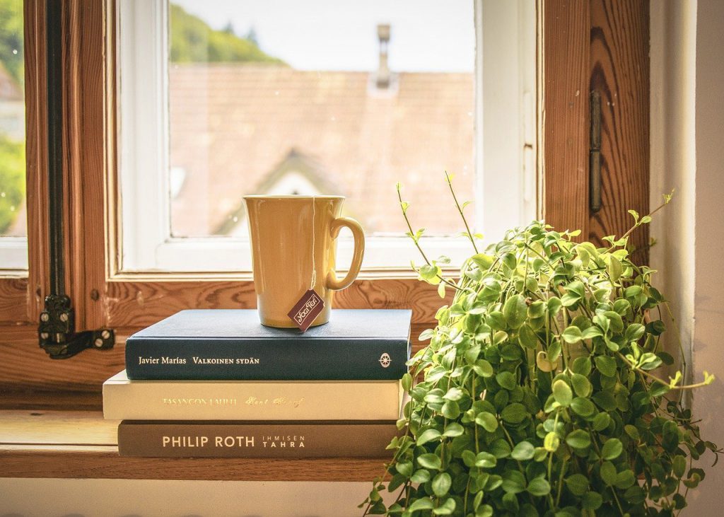 Na parapecie okiennym ułożone książki. Na książkach żółty kubek z herbatą. Obok  doniczkowy zielony kwiatek.